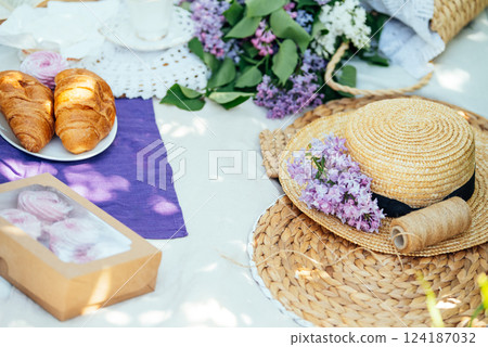 Cozy Outdoor Picnic with Croissants and Lilac Flowers charming picnic setup with croissants, tea, lilac flowers, books, and pastries on a blanket under dappled sunlight in spring day 124187032