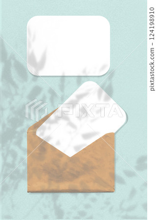 Natural light casts shadows from the leaves of a tree branch on an envelope with two sheets of textured white paper on a blue background. Mockup 124198910