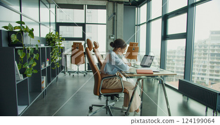 A focused professional woman working on a laptop at a modern office desk, taking notes in a notebook, surrounded by stylish furniture, plants, and natural light from large windows A focused professional woman working on a laptop at a modern office desk, taking notes in a notebook, surrounded by stylish furniture, plants, and natural light from large windows 124199064