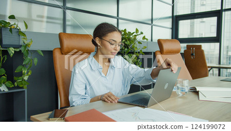 smiling young woman in glasses sitting at a modern office desk, closing her laptop while enjoying a productive day in a clean, professional, and stylish workspace smiling young woman in glasses sitting at a modern office desk, closing her laptop while enjoying a productive day in a clean, professional, and stylish workspace 124199072