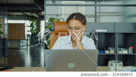 A focused young woman in glasses sitting at a modern office desk, looking at her laptop screen with a thoughtful expression in a professional and organized workspace A focused young woman in glasses sitting at a modern office desk, looking at her laptop screen with a thoughtful expression in a professional and organized workspace 124199073