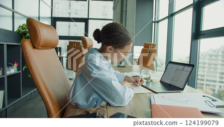 A focused professional woman working on a laptop at a modern office desk, taking notes in a notebook, surrounded by stylish furniture, plants, and natural light from large windows 124199079