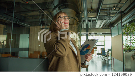 A focused businesswoman in a modern office, engaged in a phone call while holding a clipboard with data charts, showcasing confidence and professionalism A focused businesswoman in a modern office, engaged in a phone call while holding a clipboard with data charts, showcasing confidence and professionalism 124199089