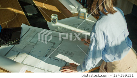 An overhead view of a professional architect analyzing blueprints at a wooden desk, accompanied by a laptop, glasses, and a glass of water in a sunlit workspace An overhead view of a professional architect analyzing blueprints at a wooden desk, accompanied by a laptop, glasses, and a glass of water in a sunlit workspace 124199131