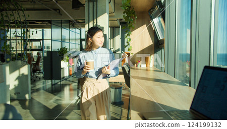 A young professional woman holding a coffee cup and clipboard in a modern office environment with large windows and natural light, emphasizing productivity and focus A young professional woman holding a coffee cup and clipboard in a modern office environment with large windows and natural light, emphasizing productivity and focus 124199132
