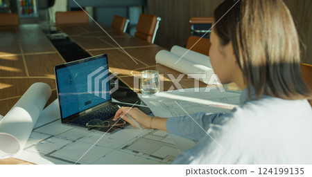 An architect reviewing detailed blueprints on a laptop in a modern conference room, surrounded by rolled plans and sunlight reflecting on a wooden table 124199135