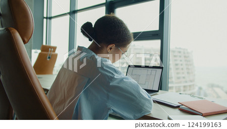 A young professional woman working on a laptop in a bright modern office space, analyzing architectural blueprints on the screen with large windows and urban views 124199163