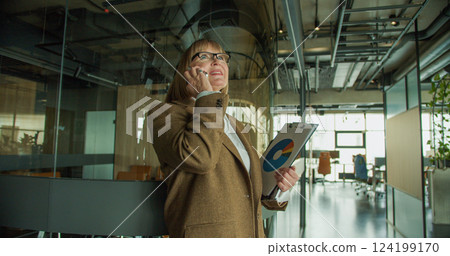 A focused businesswoman in a modern office, engaged in a phone call while holding a clipboard with data charts, showcasing confidence and professionalism A focused businesswoman in a modern office, engaged in a phone call while holding a clipboard with data charts, showcasing confidence and professionalism 124199170