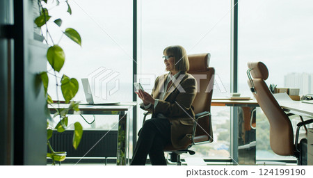 A focused businesswoman using smartphone while sitting in a modern office with a large window view overlooking the city and greenery A focused businesswoman using smartphone while sitting in a modern office with a large window view overlooking the city and greenery 124199190