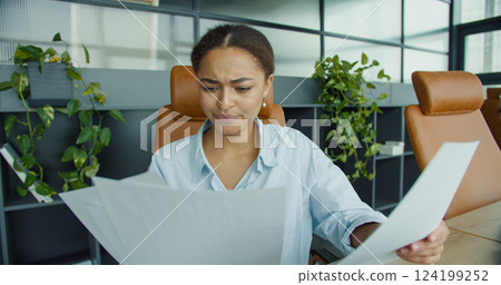 focused young woman analyzing documents at her desk in a modern office, surrounded by charts, a laptop, and office supplies with stylish leather chairs 124199252