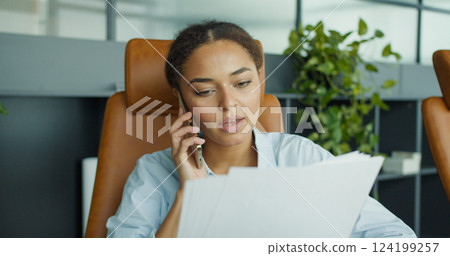 A young professional woman talking on the phone while reviewing documents in a modern office with plants and stylish leather chairs in the background A young professional woman talking on the phone while reviewing documents in a modern office with plants and stylish leather chairs in the background 124199257