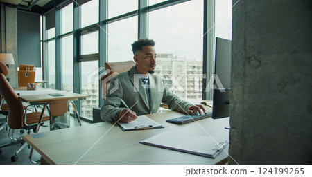 A professional businessman working at his desk in a modern office with large windows, balancing note-taking and computer work in a sleek corporate environment. A professional businessman working at his desk in a modern office with large windows, balancing note-taking and computer work in a sleek corporate environment. 124199265