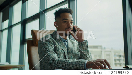 A professional businessman in a modern office taking a phone call while working, showcasing multitasking and productivity in a corporate environment. 124199280