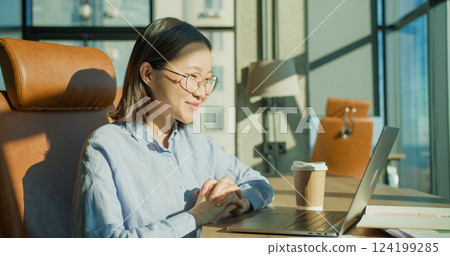 A young woman relaxing with her hands behind her head in a modern office with a laptop and coffee cup on the desk, enjoying sunlight through large windows 124199285
