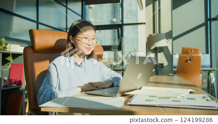 A cheerful woman in glasses waving during a video call in a modern sunlit office with a laptop, coffee cup, and stylish furniture creating a productive atmosphere 124199286