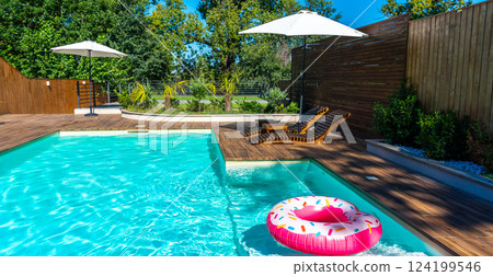 Modern backyard swimming pool with turquoise water, wooden deck, umbrellas, lounge chairs, and pink donut inflatable Modern backyard swimming pool with turquoise water, wooden deck, umbrellas, lounge chairs, and pink donut inflatable 124199546