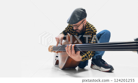 Focused man, musician crouching, playing double bass, deeply immersed in rhythm on minimalist white backdrop. Focused man, musician crouching, playing double bass, deeply immersed in rhythm on minimalist white backdrop. 124201255