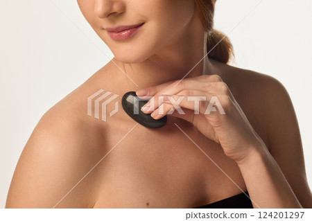 Close-up of woman massaging shoulder with black stone for deep relaxation and glowing skin on white background. 124201297