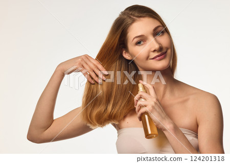 Beautiful young woman taking care after her hair, applying moisturizing and protective hair spray against white background 124201318