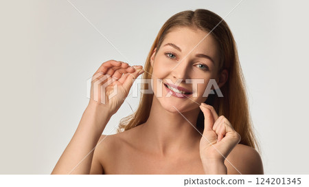 Smiling young woman with radiant skin using dental floss, emphasizing healthy teeth and oral hygiene in soft natural lighting 124201345