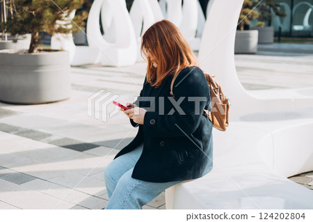 Happy woman chatting on phone and relaxing outdoors. Urban, people concept. Smiling 30s redhead woman text messaging on smart phone at sunny day on the city street, spring time 124202804