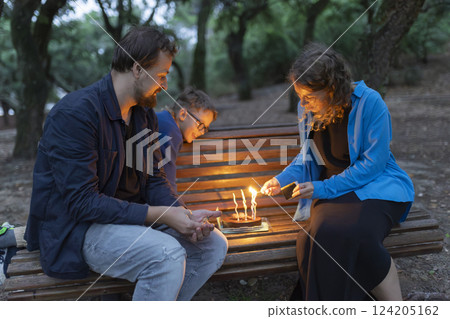 Family celebrating birthday with cake and candles in park at night 124205162