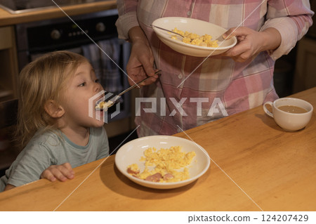 A woman feeds her child scrambled eggs in the kitchen. 124207429