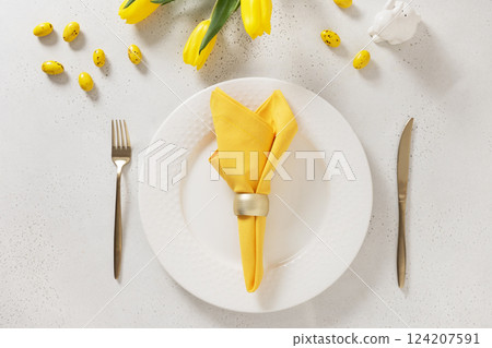 Easter table setting with spring tulips, white plates, bunny, eggs and yellow decoration on white background. Happy Easter celebration. Copy space. View from above. 124207591
