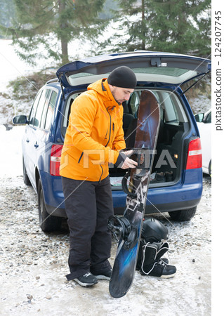 Preparing for an adventurous day of snowboarding in a snowy mountain landscape while enjoying the crisp winter air and scenic views alongside a parked blue vehicle Preparing for an adventurous day of snowboarding in a snowy mountain landscape while enjoying the crisp winter air and scenic views alongside a parked blue vehicle 124207745