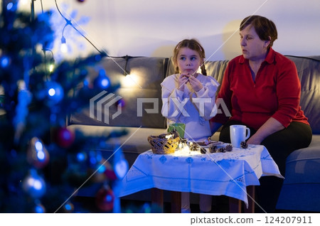 Cozy Christmas evening: A grandmother and granddaughter share treats and warm drinks by a decorated tree. 124207911