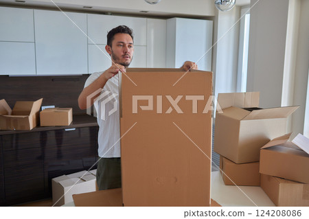 Man standing in living room on day of move to rented home, unbox carton box with belongings. Relocate to new own apartment collect personal things Man standing in living room on day of move to rented home, unbox carton box with belongings. Relocate to new own apartment collect personal things 124208086