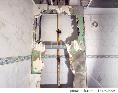 A detailed closeup view of a wall that has been significantly damaged, inside it a visible pipe with rust at plumbing joint and broken tiles 124208096
