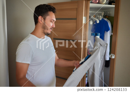 Domestic Device. Happy Middle Eastern Young Man Using Clothes Steamer And Steaming Shirt, Ironing His Clothes With Portable Gadget Domestic Device. Happy Middle Eastern Young Man Using Clothes Steamer And Steaming Shirt, Ironing His Clothes With Portable Gadget 124208133