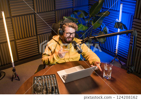 Cheerful young man recording podcast in studio. Confident man with headphones and microphone recording a podcast. Happy guy talking on web radio. Cheerful young man recording podcast in studio. Confident man with headphones and microphone recording a podcast. Happy guy talking on web radio. 124208162