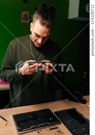 Vertical portrait of professional technician carefully examining small component from laptop motherboard, troubleshooting and performing repairs in specialized workshop with green background. 124208712