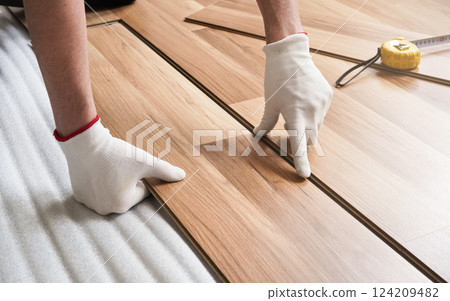 Installing laminated floor, detail on man hands in gloves fitting wooden tile, over white foam base layer, tape measure in background Installing laminated floor, detail on man hands in gloves fitting wooden tile, over white foam base layer, tape measure in background 124209482