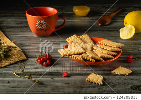 A cozy setup with a bowl of crispy biscuits, a cup of herbal tea, sliced lemon, and some honey on a rustic wooden table. Fresh herbs add a touch of nature to this snack moment 124209813