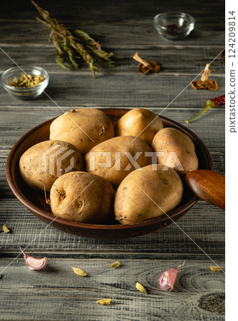 A wooden bowl contains several freshly harvested potatoes, surrounded by various spices and garlic on a textured rustic table, creating a warm culinary atmosphere 124209814