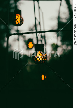 A decorative garden garland with black lanterns and burning bulbs on a garden arch against a dark evening sky. Bulb lamps in twilight. Festive decor. 124210312