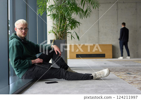 A casual young professional sitting by a large window in a modern office, smiling while working on a laptop, with colleagues interacting in the blurred background. A casual young professional sitting by a large window in a modern office, smiling while working on a laptop, with colleagues interacting in the blurred background. 124211997