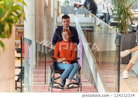 A company director assists his business colleague in a wheelchair, helping her navigate to their startup office, where they work alongside their diverse team of colleagues, emphasizing inclusivity and A company director assists his business colleague in a wheelchair, helping her navigate to their startup office, where they work alongside their diverse team of colleagues, emphasizing inclusivity and 124212154