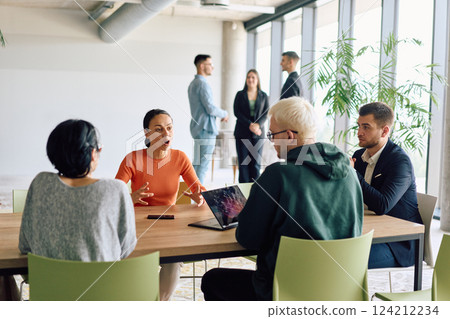 A diverse team of business professionals engaged in a discussion around a conference table in an office, while their colleagues collaborate in the background 124212234