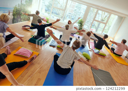 A group of senior women engage in various yoga exercises, including neck, back, and leg stretches, under the guidance of a trainer in a sunlit space, promoting well-being and harmony 124212299