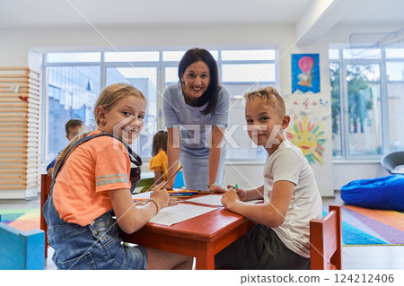 Creative kids during an art class in a daycare center or elementary school classroom drawing with female teacher. 124212406