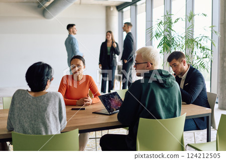 A diverse team of business professionals engaged in a discussion around a conference table in an office, while their colleagues collaborate in the background A diverse team of business professionals engaged in a discussion around a conference table in an office, while their colleagues collaborate in the background 124212505