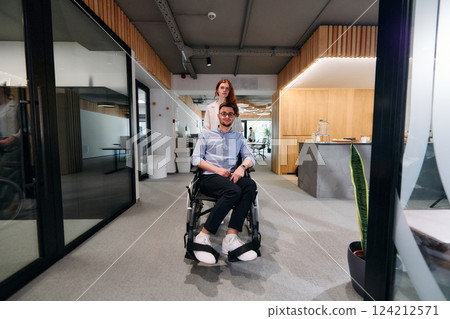 Business colleagues, collaborative business colleagues, including a person in a wheelchair, walk past a modern glass office corridor, illustrating diversity, teamwork and empowerment in the workplace. Business colleagues, collaborative business colleagues, including a person in a wheelchair, walk past a modern glass office corridor, illustrating diversity, teamwork and empowerment in the workplace. 124212571