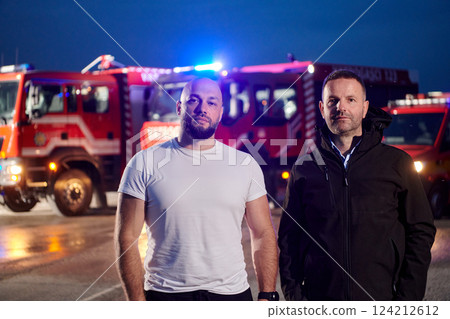 Group of firefighters, dressed in civilian clothing, stand in front of fire trucks during the night, showcasing a moment of camaraderie and unity among the team as they reflect on their duties and the 124212612