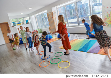 Small nursery school children with female teacher on floor indoors in classroom, doing exercise. Jumping over hula hoop circles track on the floor. Small nursery school children with female teacher on floor indoors in classroom, doing exercise. Jumping over hula hoop circles track on the floor. 124212631
