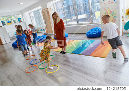 Small nursery school children with female teacher on floor indoors in classroom, doing exercise. Jumping over hula hoop circles track on the floor. 124212705