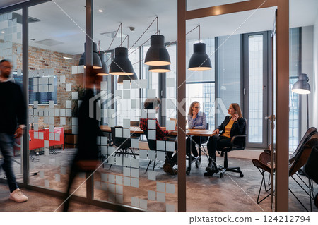In the dynamic hustle and bustle of a business environment, a group of young business professionals walk down the corridor next to their office, while their colleagues collaborate inside, symbolizing 124212794